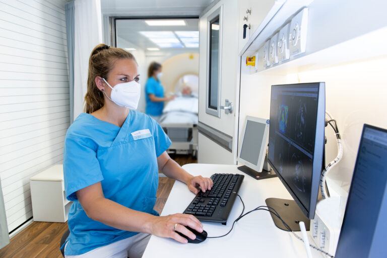 Medizinische Fachkraft in blauer Schutzkleidung und Maske bedient Computer mit medizinischen Bildgebungen, im Hintergrund Patient auf Liege und weitere Fachkraft in Krankenzimmer.