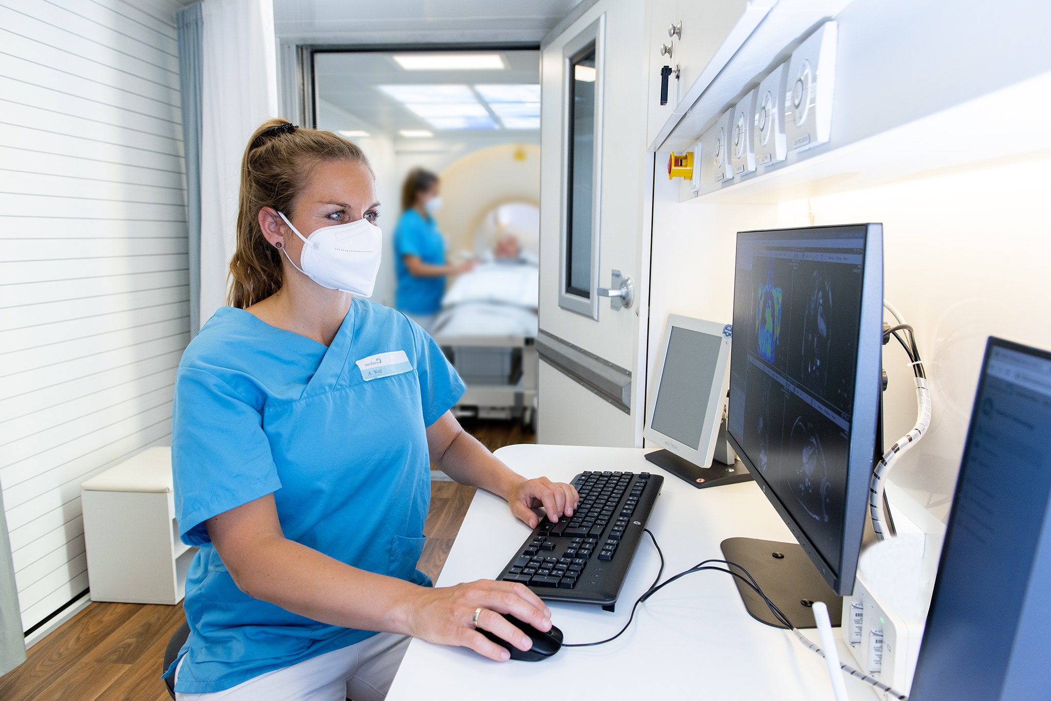 Medizinische Fachkraft in blauer Schutzkleidung und Maske bedient Computer mit medizinischen Bildgebungen, im Hintergrund Patient auf Liege und weitere Fachkraft in Krankenzimmer.
