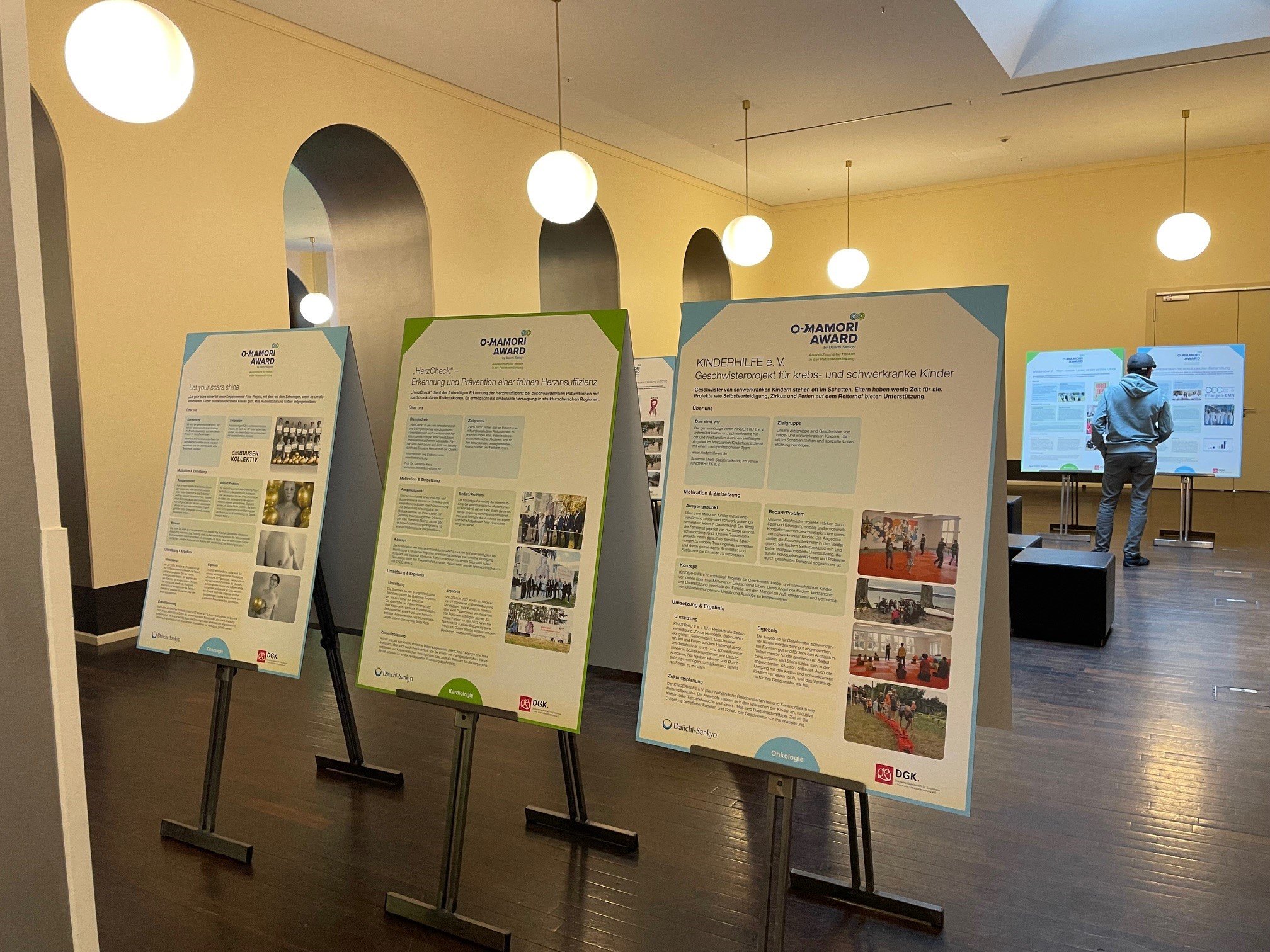 Ausstellung mit drei Informationsplakaten auf Staffeleien in einem hellen Raum mit Holzboden und runden Hängeleuchten, im Hintergrund ein weiterer Raum mit zwei zusätzlichen Plakaten, vor denen eine Person in grauer Kleidung steht.
