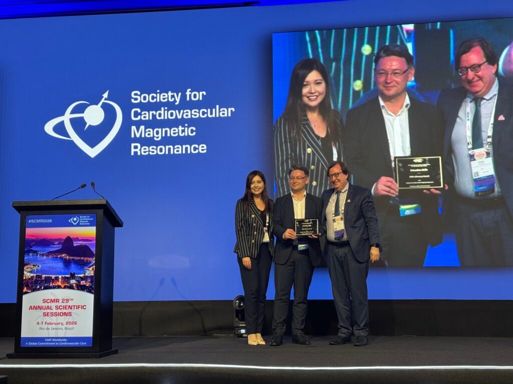 Drei Personen stehen auf einer Bühne mit einem Schild der Society for Cardiovascular Magnetic Resonance. Zwei Personen halten eine Auszeichnung in der Hand. Eine große Leinwand im Hintergrund zeigt das gleiche Bild.