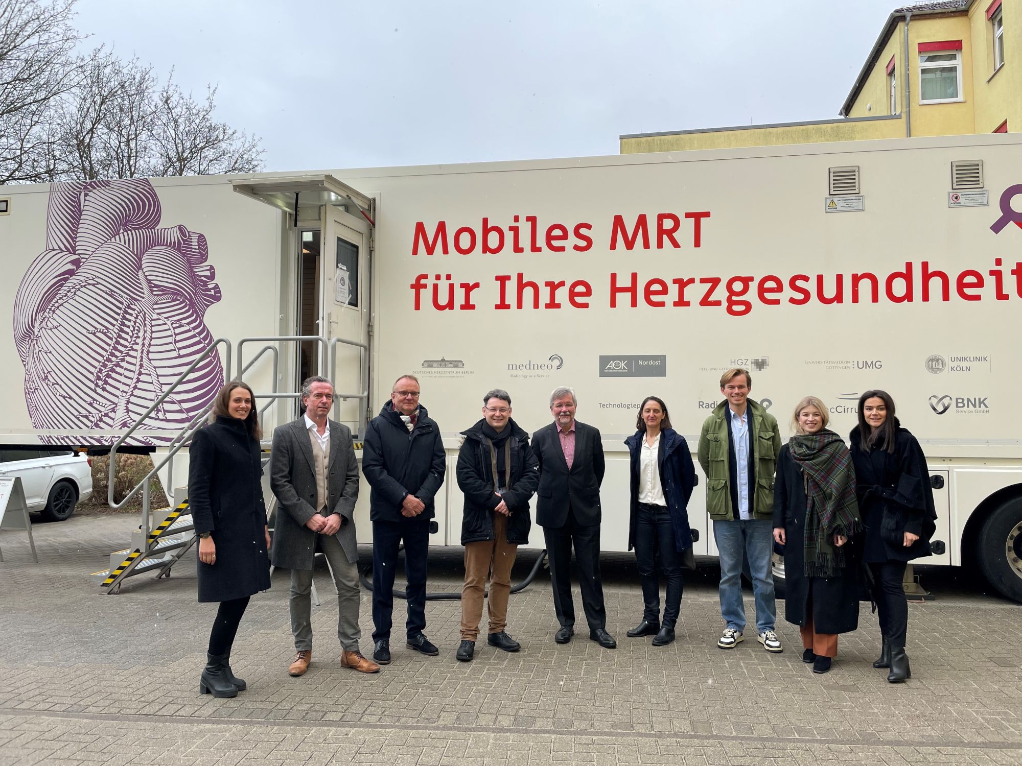 Neun Personen stehen vor einem weißen mobilen MRT-Bus mit der Aufschrift "Mobiles MRT für Ihre Herzgesundheit" und einer großen grafischen Abbildung eines Herzens, die Tür des Busses ist geöffnet.