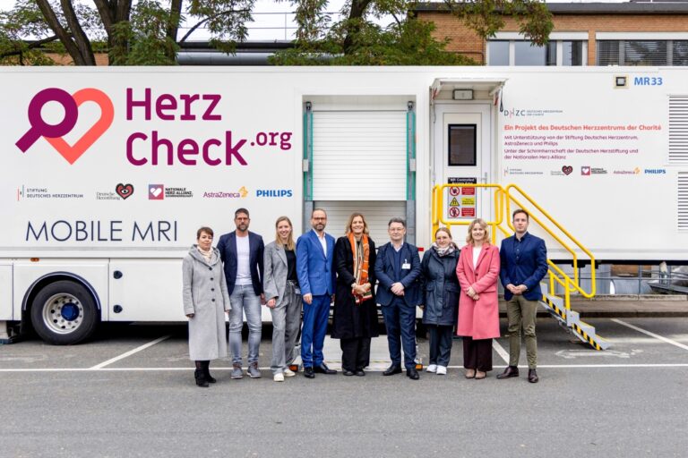 Eine Gruppe von zehn Personen steht vor einem mobilen Herzdiagnosefahrzeug mit der Aufschrift HerzCheck.org und Mobile MRI. Es ist ein Projekt des Deutschen Herzzentrums, unterstützt von mehreren Organisationen.