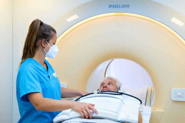 Krankenpflegerin in blauem Kittel und medizinischer Maske bereitet männlichen Patienten auf eine MRT-Untersuchung in einem Philips-Medizingerät vor, Patient liegt auf Untersuchungsliege und blickt nach oben.
