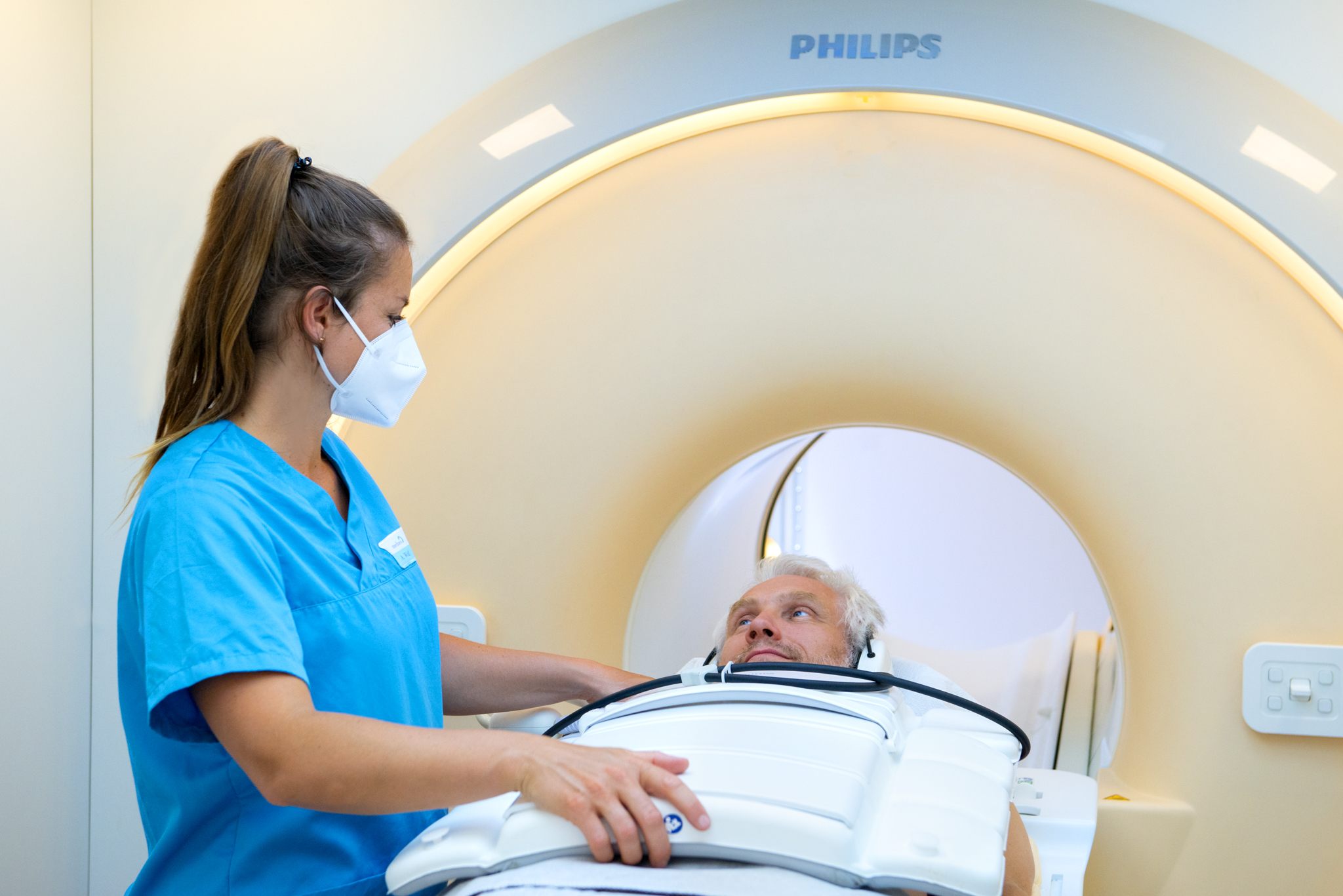 Krankenpflegerin in blauem Kittel und medizinischer Maske bereitet männlichen Patienten auf eine MRT-Untersuchung in einem Philips-Medizingerät vor, Patient liegt auf Untersuchungsliege und blickt nach oben.