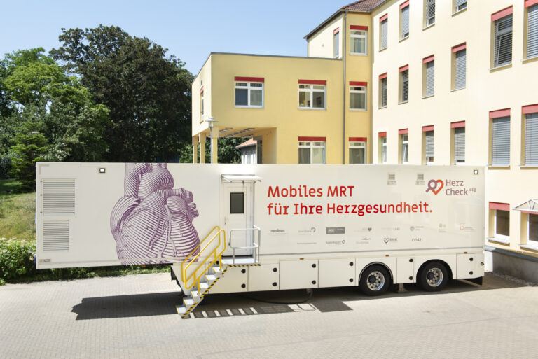 Mobilitätstauglicher MRT-Wagen mit gelb lackierter Außentreppe, bedruckt mit einem großen stilisierten menschlichen Herzen und dem Schriftzug 'Mobiles MRT für Ihre Herzgesundheit' vor gelb gestrichenem Gebäudekomplex mit roten Fensterrahmen und grünem Baumbestand im Hintergrund