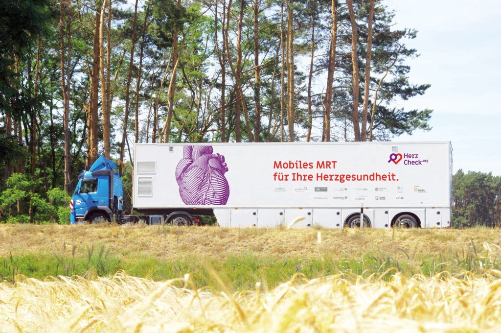Blauer LKW mit einem weißen Anhänger, der Aufschrift "Mobiles MRT für Ihre Herzgesundheit" und einer großen lila Grafik eines Herzens, parkend vor einem Wald mit grünen Bäumen im Hintergrund, auf einer grasbewachsenen Fläche bei Sonnenschein.