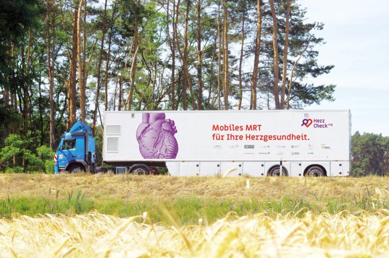 Blauer LKW mit einem weißen Anhänger, der Aufschrift "Mobiles MRT für Ihre Herzgesundheit" und einer großen lila Grafik eines Herzens, parkend vor einem Wald mit grünen Bäumen im Hintergrund, auf einer grasbewachsenen Fläche bei Sonnenschein.