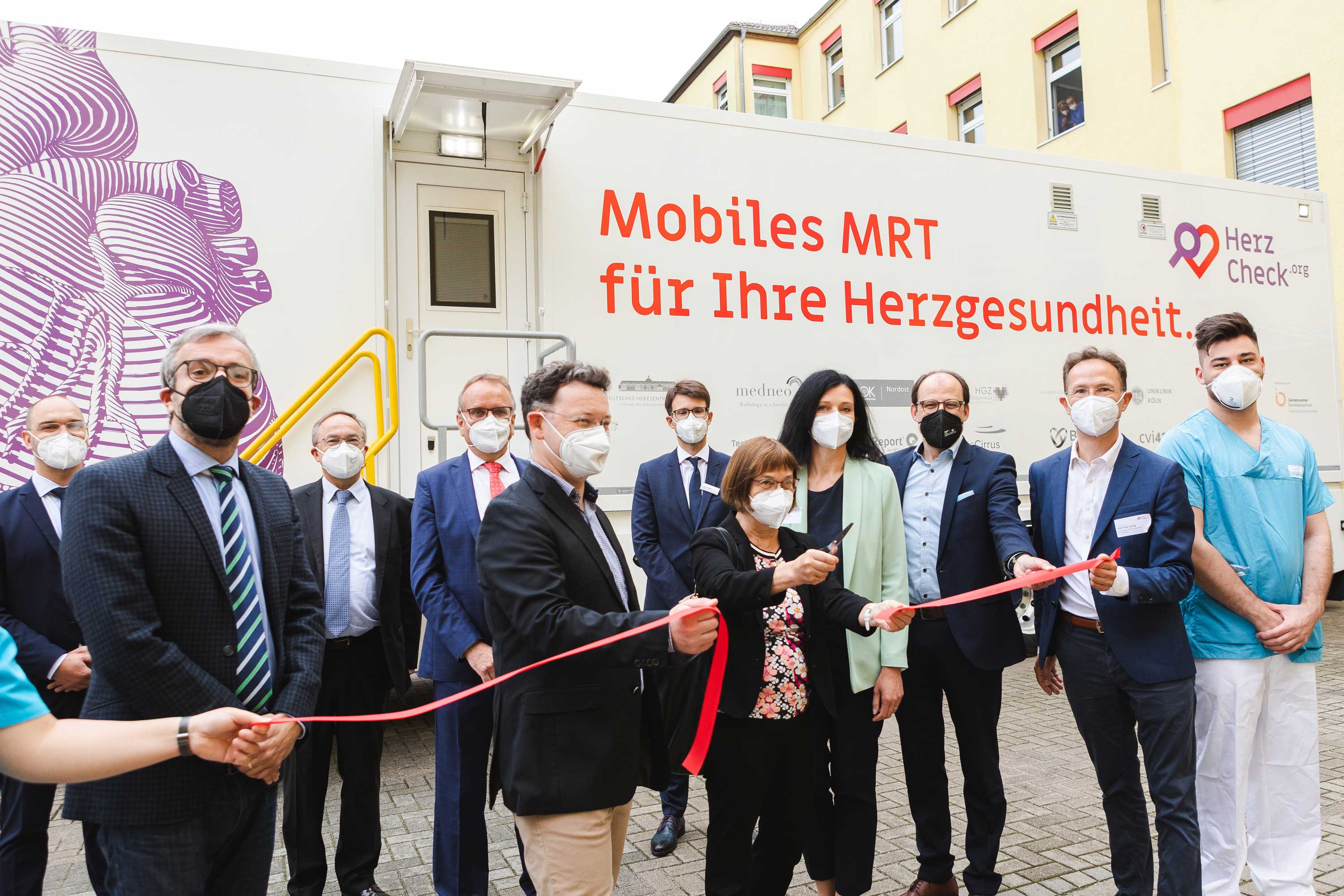 Gruppe von Menschen mit Masken bei der Eröffnung eines mobilen MRT-Systems zur Herzgesundheit, Personen schneiden ein rotes Band vor einem weißen Fahrzeug mit der Aufschrift "Mobiles MRT für Ihre Herzgesundheit" und einem großen lila Herzgrafik.